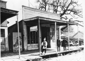 false front wooden building with man standing under sign reading Sung Lung Washing and Ironing. Man stands on wooden sidewalk, dressed in clothing typical for Chinese men of the time period.