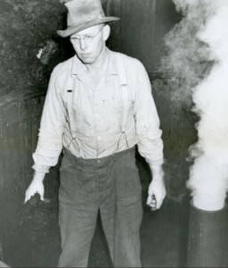Man standing in dye vat next to steaming ppe with wool in hands and hat on head.