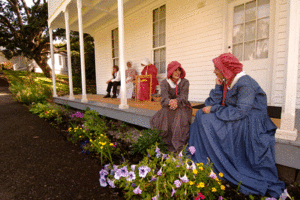 White historical home front porch with several people dressed in period clothing conversing.