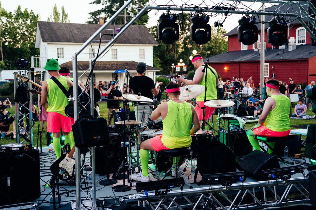 view of band at Make Music Day dressed in bright neon colors from behind on stage at Willamette Heritage Center. Audience, historical house, and warehouse in background.